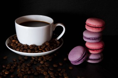 Siyah kokulu kahveli beyaz bir kupa siyah bir zurnanın üzerinde renkli Fransız kurabiyelerinin yanında duruyor.