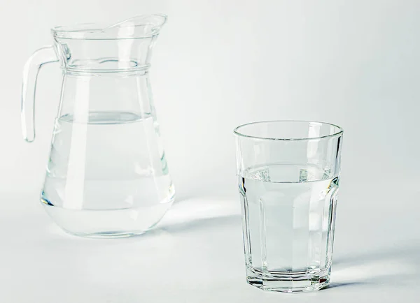 Acqua pura e limpida in un bicchiere di vetro e una brocca di vetro poggia su uno sfondo bianco — Foto Stock