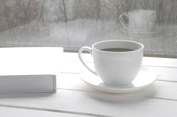 Maison confortable nature morte : tasse blanche de café chaud et livre blanc ouvert sur le rebord de la fenêtre contre le paysage de neige à l'extérieur . — Photo