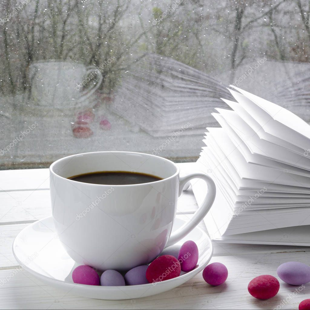 Cozy concept: a white cup of coffee stands on a white wooden table next to a white open book and colorful candy against the background of a window with raindrops