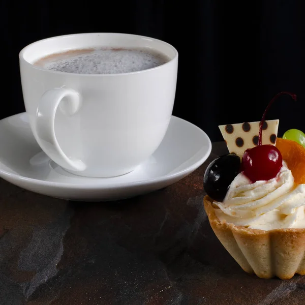 Bolo de frutas com cerejas, uvas e ameixa está em uma pedra selvagem ao lado de uma xícara de café branco com uma espuma — Fotografia de Stock