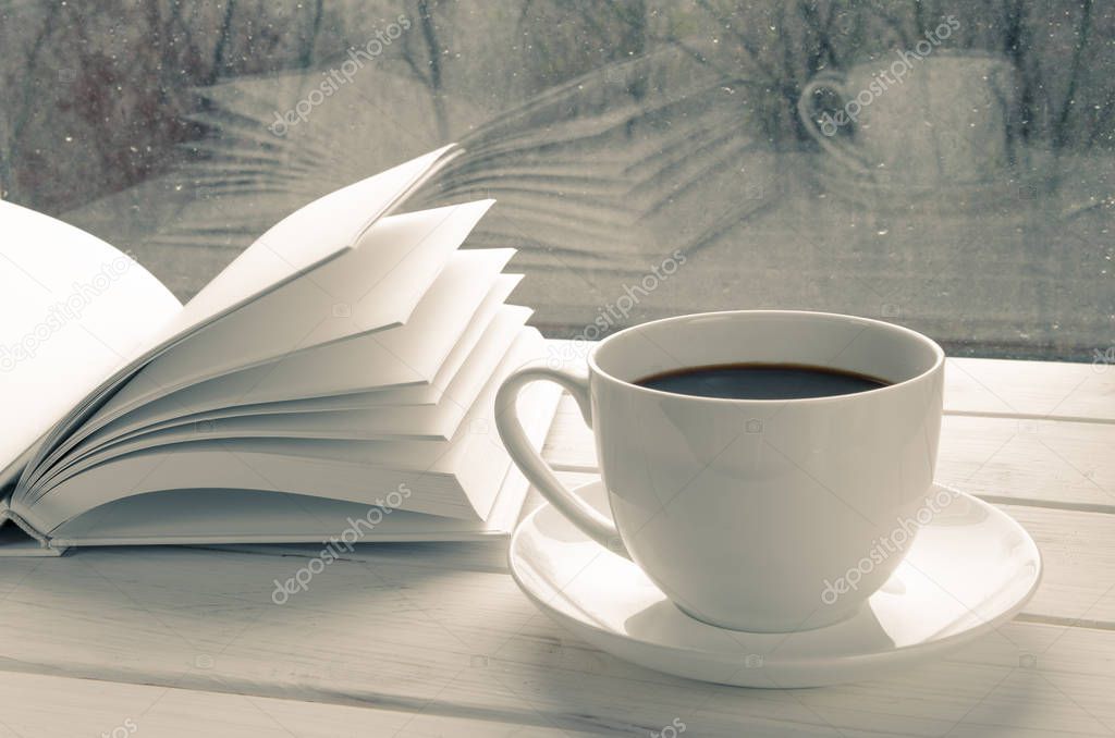 Cozy home still life: white cup of hot coffee and white opened book on windowsill against snow landscape outside.