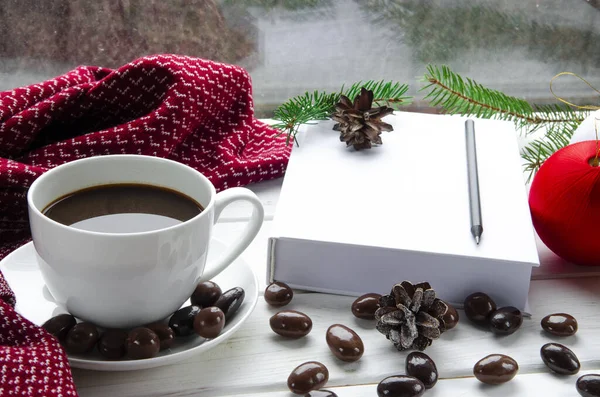 Concepto de Navidad: una taza de café blanco se coloca sobre una mesa de madera blanca junto a un libro blanco abierto y dulces de colores sobre el fondo de una ventana con gotas de lluvia — Foto de Stock