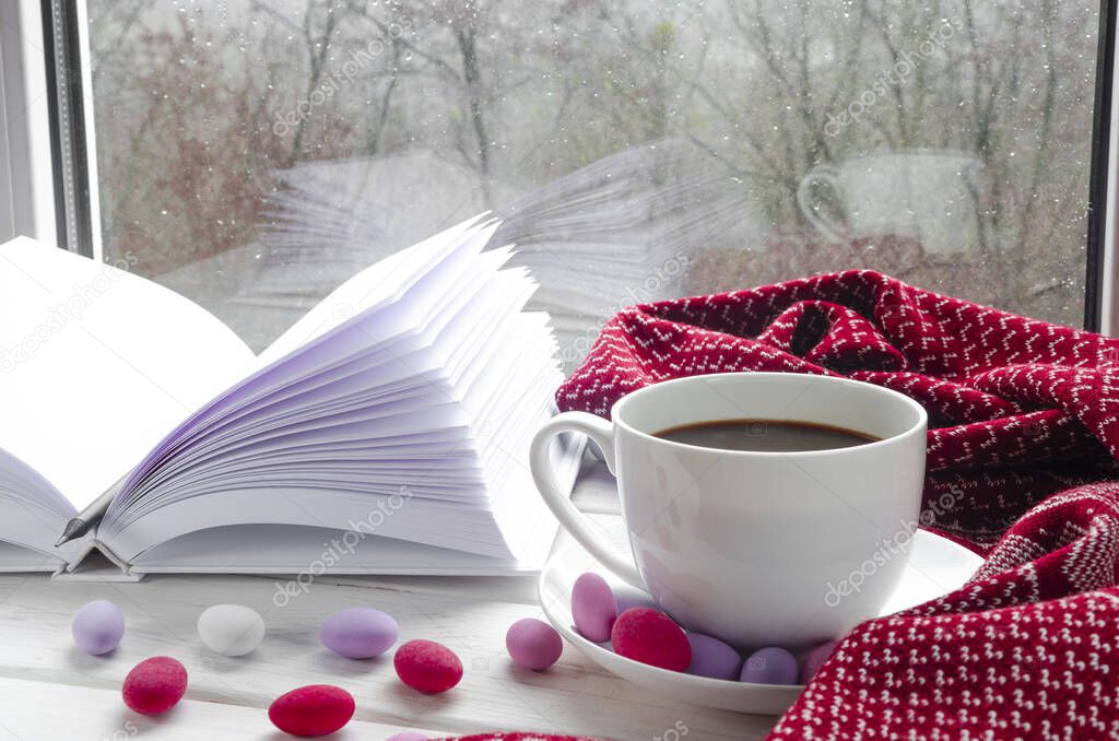 Cozy home still life: cup of hot coffee and opened book with warm plaid on windowsill against snow landscape outside