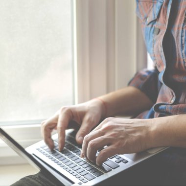 Bir adam elinde dizüstü bilgisayarla pencere eşiğinde oturur ve çalışır.