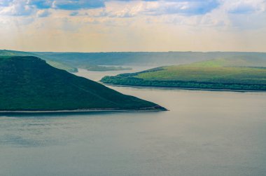 Yeşil ağaçlarla kaplı dağlar mavi gökyüzüne karşı bir dağ gölüne iniyor.