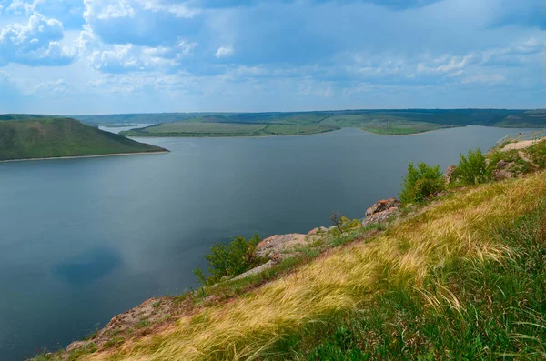 Yeşil ağaçlarla kaplı dağlar mavi gökyüzüne karşı bir dağ gölüne iniyor.