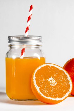Taze sıkılmış portakal suyuyla dolu cam kavanoz taze portakalların yanında beyaz arka planda duruyor.
