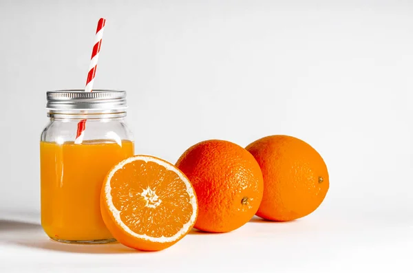 Tarro de vidrio con un tubo con zumo de naranja recién exprimido sobre un fondo blanco junto a naranjas frescas —  Fotos de Stock