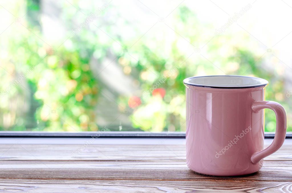 A pink cup of tea stands on a wooden window sill against the background of a summer garden