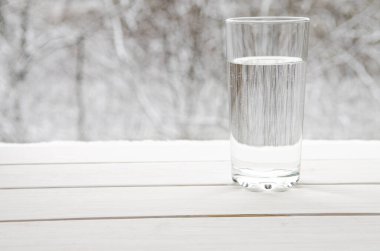 A glass of transparent melt water stands on a white wooden window sill against the backdrop of winter snow-covered forest
