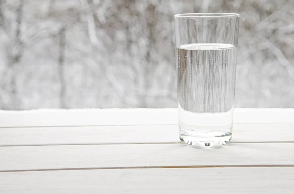 A glass of transparent melt water stands on a white wooden window sill against the backdrop of winter snow-covered forest — Stock Photo, Image