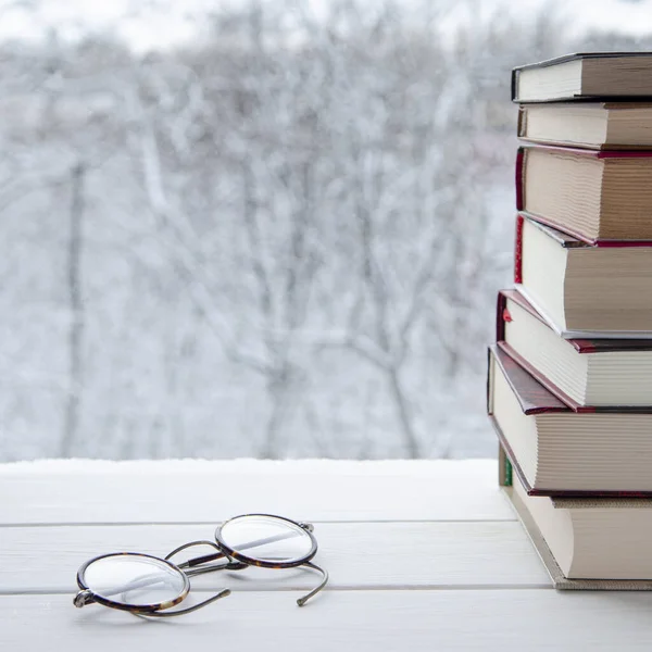 Old multi-colored books stand on a wooden shelf against the backdrop of the winter forest