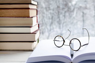 Yuvarlak okuma gözlükleri, açık bir kitabın üzerinde, beyaz sayfaların yanında, ahşap bir pencere pervazında, çok renkli kitapların yanında.