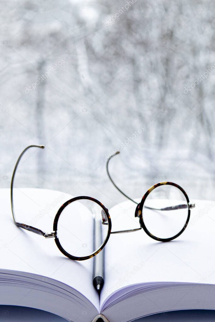 Round glasses and a black pencil lie on a notepad with white pages against the background of the landscape outside the window. Wall Background