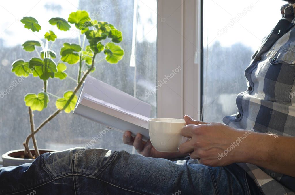 A man sits on a window sill with a cup of coffee and a book during quarantine due to the coronavirus pandemic