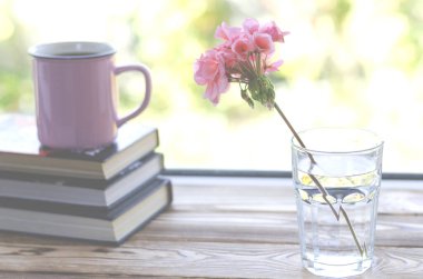 Pembe bir sardunya çiçeği bir bardak temiz suyun içinde duruyor tahta bir pencere eşiğinde kitaplar ve pembe bir fincan çayın yanında.