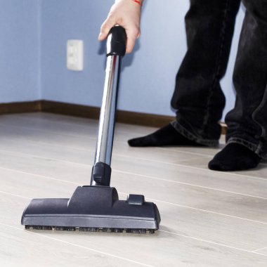 Elinde elektrikli süpürge olan bir adam mavi duvarlı ve ışıklı zemini olan bir odada temizlik yapıyor.