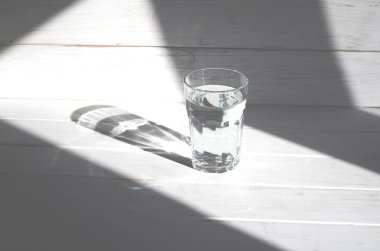 Keskin gölgeleri olan beyaz ahşap bir zeminde bir bardak temiz temiz su duruyor.