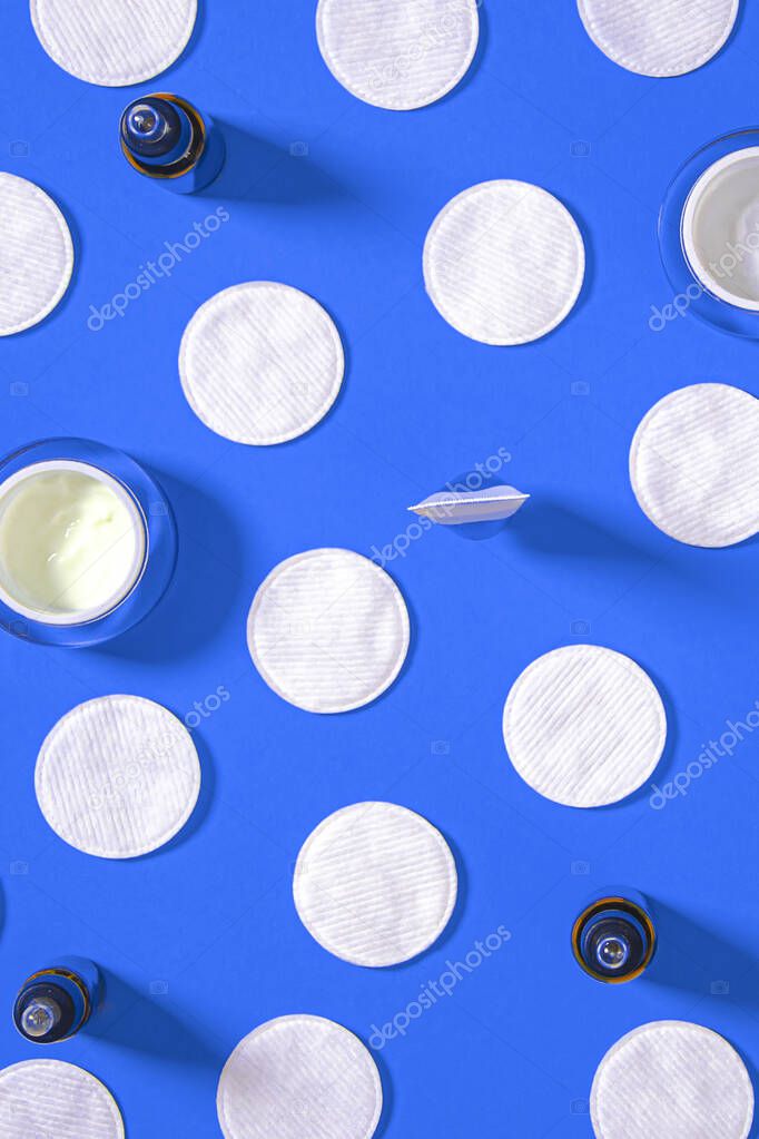 Hygiene products: white round cotton pads and plastic sticks for cleaning the ears lie on a blue background next to face cream. Top view, flat lay
