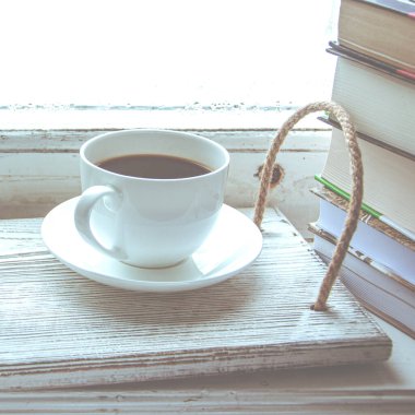 Beyaz bir fincan kahve ve bir bardak su sehpası pencere kenarındaki ahşap tepside bir yığın kitabın yanında. Yakın plan.