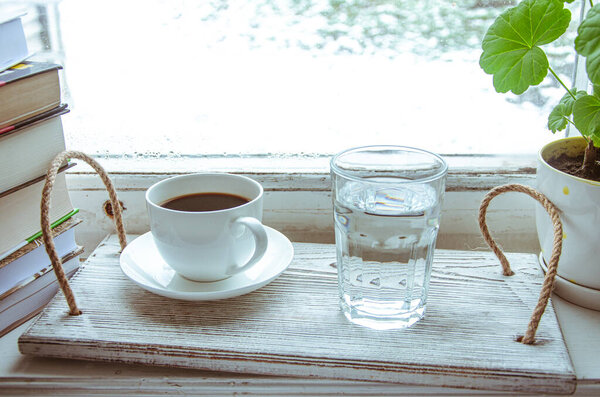 A white cup of coffee and a glass of water stand on a wooden tray on the windowsill next to a stack of books. Close-up