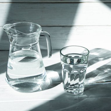 Keskin gölgeleri olan beyaz ahşap bir zeminde bir bardak temiz temiz su duruyor. gölgeler