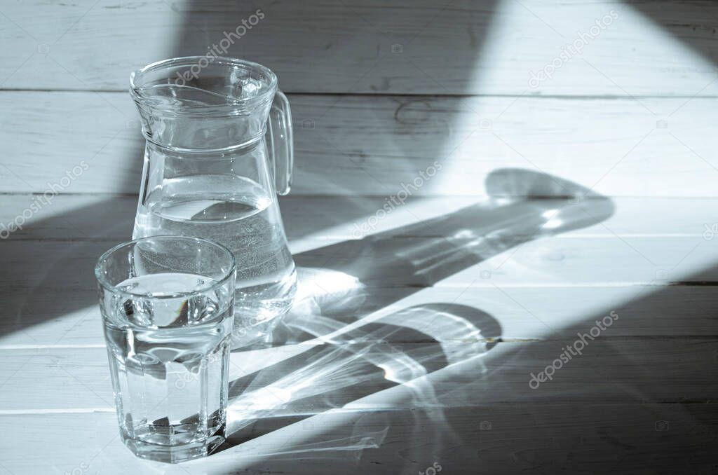 Sharp shadows from the carafe and a glass of water on a white wooden background