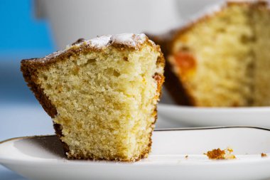 Ön planda, kesilmiş bir parça tatlı kek beyaz bir fincan tabağının üzerinde duruyor. Arka planda kekin geri kalanı ve mavi arkaplandaki beyaz bardak yer almıyor..
