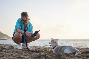 Genç, güzel, sarışın bir kadın, telefonuyla yavru köpeğinin fotoğrafını çekiyor. Sahibi ve açık gri köpeği, gün batımında kumlu sahilde oynuyorlar..
