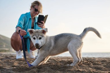 Genç, güzel, sarışın bir kadın, telefonuyla yavru köpeğinin fotoğrafını çekiyor. Sahibi ve açık gri köpeği, gün batımında kumlu sahilde oynuyorlar..