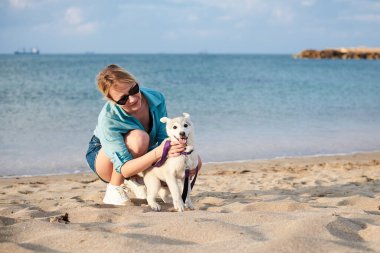 Sahilde sahibinin ellerinde tutulan küçük, açık gri bir köpek yavrusu. Genç bir kadın, turkuaz bluz, kot şort ve beyaz spor ayakkabı giyiyor, köpeğinin ağzını tutuyor,.