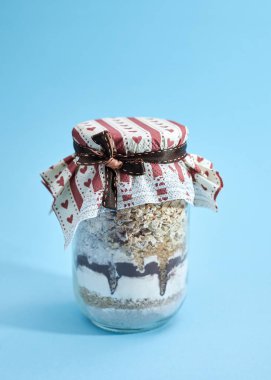 Mısır gevreği, un, kahverengi ve beyaz şeker ve çikolata tozu katmanları, kahverengi peçete ile kaplı cam kavanoz ve açık mavi arkaplan üzerine fiyonk