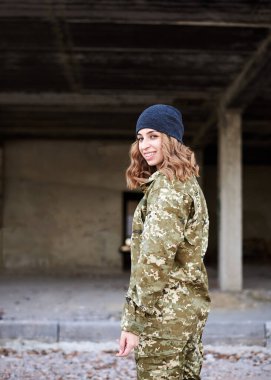 Genç kıvırcık saçlı asker kadın, Ukrayna askeri üniforması ve koyu gri şapka giyiyor, resim için poz veriyor. Harap olmuş bir binanın önündeki askerin çeyrek portresi. İnşaat alanı..