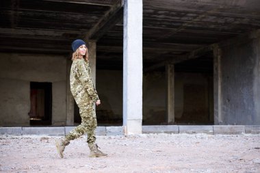 Kıvırcık saçlı asker kadın, Ukrayna askeri üniforması giyiyor ve koyu gri şapkası var, gülümsüyor. Harap olmuş inşaat alanının önünde yürüyen asker portresi..