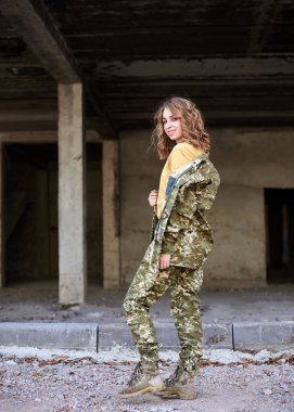 Genç kıvırcık saçlı asker kadın, Ukrayna askeri üniforması giyiyor, resim için poz veriyor. Harap olmuş bir binanın önündeki asker portresi. İnşaat alanı..