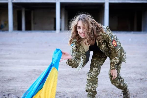 若い巻きブロンドの軍の女性は ウクライナ軍の制服を着て 空気中のウクライナの青と黄色の旗を投げます 廃墟と化した建物の前の女性兵士の四分の一の肖像画 — ストック写真