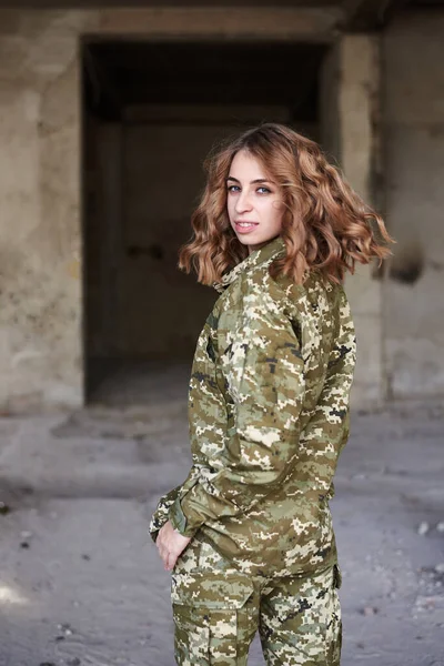 Young curly blond military woman, wearing ukrainian military uniform, posing for picture. Three-quarter portrait of soldier in front of ruined abandoned building, construction site.