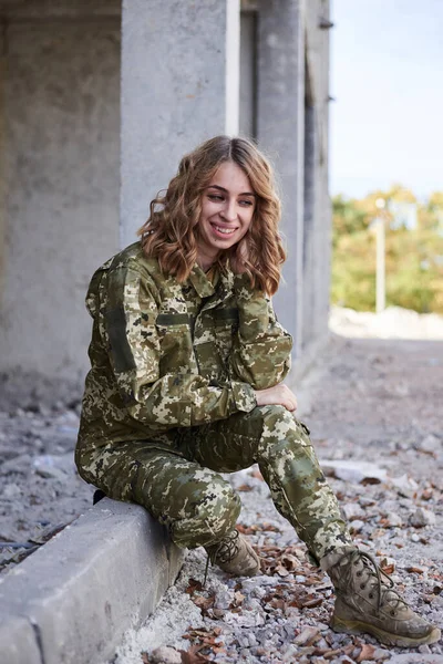 Young curly blond military woman, wearing ukrainian military uniform, sitting on pavement edge, smiling. Full-length portrait of army soldier in front of ruined abandoned building, construction site.