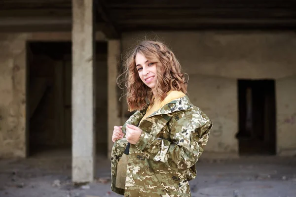 Young curly blond military woman, wearing ukrainian military uniform, posing for picture. Three-quarter portrait of soldier in front of ruined abandoned building, construction site.