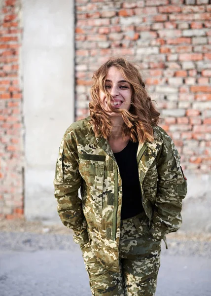 Young curly blond military woman, wearing ukrainian military uniform, posing for picture. Three-quarter portrait of female army soldier standing in front of brick wall of ruined building, smiling.