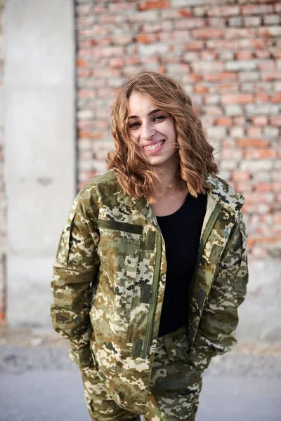 Young curly blond military woman, wearing ukrainian military uniform, posing for picture. Three-quarter portrait of female army soldier standing in front of brick wall of ruined building, smiling.