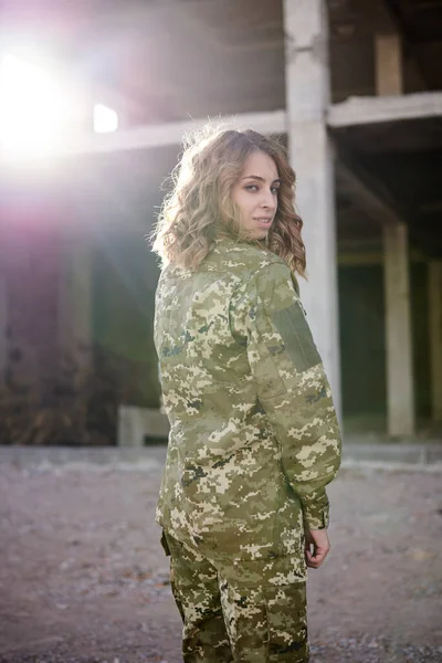 Young curly blond military woman, wearing ukrainian army military uniform and black t-shirt. Three-quarter portrait of female soldier in rim light glare in front of ruined abandoned building.