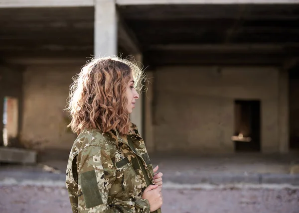 Young curly blond military woman, wearing ukrainian army military uniform and black t-shirt. Three-quarter portrait of female soldier in rim light glare in front of ruined abandoned building.