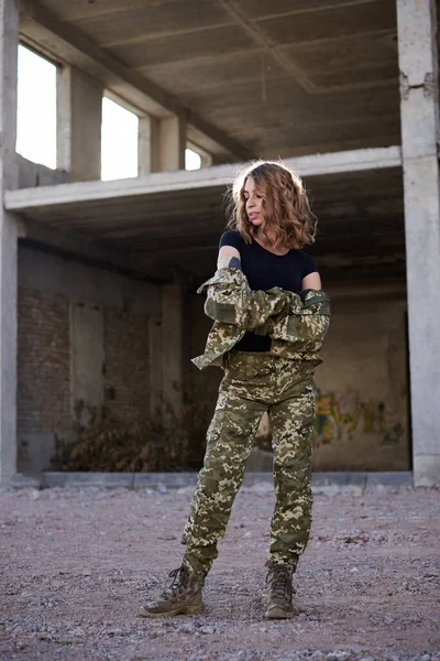 Young curly blond military woman, wearing ukrainian army military uniform and black t-shirt. Full-length portrait of female soldier standing in rim light glare in front of ruined abandoned building.