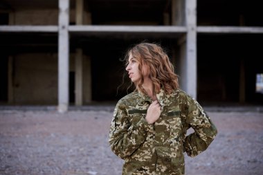 Kıvırcık saçlı asker kadın, Ukrayna askeri üniforması ve siyah tişört giyiyor, poz veriyor. Harap olmuş bir binanın önündeki kadın askerin çeyrek portresi..