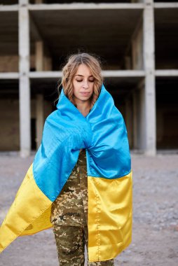 Ukrayna askeri üniforması giyen, Ukrayna mavisi ve sarı bayrağı olan kıvırcık saçlı, genç bir asker. Yıkılmış bir binanın önündeki kadın askerin çeyrek portresi..