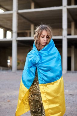 Ukrayna askeri üniforması giyen, Ukrayna mavisi ve sarı bayrağı olan kıvırcık saçlı, genç bir asker. Yıkılmış bir binanın önündeki kadın askerin çeyrek portresi..