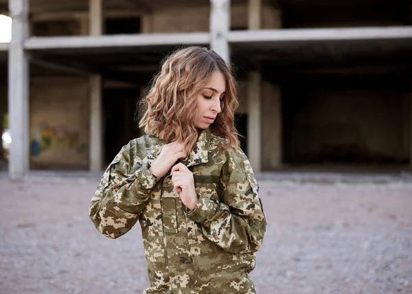 Young curly blond military woman, wearing ukrainian army military uniform and black t-shirt, holding her collar. Three-quarter portrait of female soldier in front of ruined abandoned building.