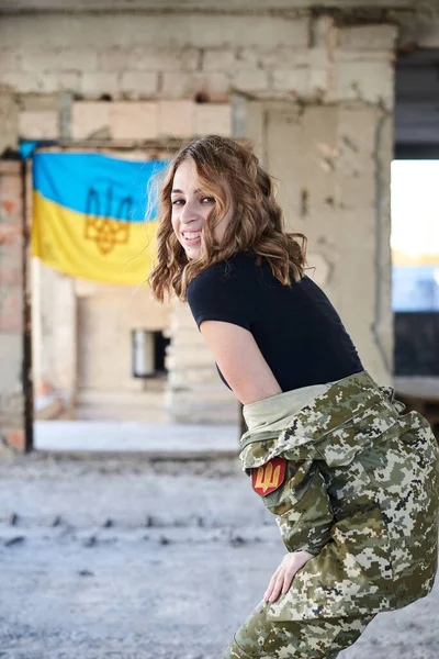 Young curly blond military woman,wearing ukrainian army uniform and black t-shirt. Three-quarter portrait of female soldier inside abandoned building with ukrainian blue and yellow flag at the back.